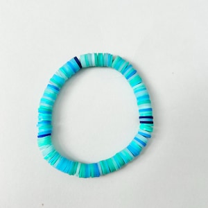 Puede incluir: Una pulsera de cuentas en forma de disco en tonos azules y turquesas. Las cuentas están dispuestas en forma circular, con una mezcla de azules y verdes claros y oscuros. Las cuentas son de tamaño y forma uniformes, creando un aspecto suave y cohesivo.