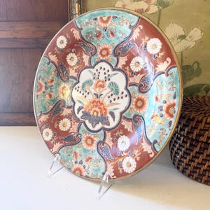 May include: A decorative ceramic plate with a floral design. The plate features a central white floral medallion surrounded by a ring of orange, teal, and brown floral patterns. The plate has a gold rim and is displayed on a clear stand.