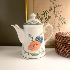 May include: White ceramic teapot with a lid, featuring a floral design with orange and blue poppies and green leaves. The teapot has a curved spout and handle, and is trimmed with green lines. A decorative piece for the kitchen or dining room.