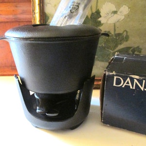 May include: A black cast iron fondue pot with a lid and handles, sitting on a matching stand. The pot is next to a black box with the word "DANSK" printed on it. The fondue pot is designed for melting cheese or chocolate.
