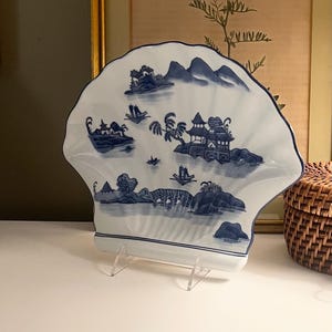 May include: A white ceramic decorative plate shaped like a large scallop shell. The surface features a blue transferware-style pattern depicting a landscape with pagodas, small boats on water, a bridge, trees, and distant mountains.