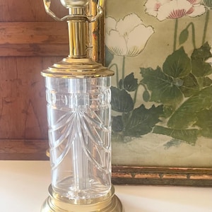 May include: A table lamp featuring a clear, cylindrical glass base with vertical etched detailing and a scalloped pattern. The lamp has a polished brass base, a matching brass neck, and a metal socket assembly at the top for a light bulb.