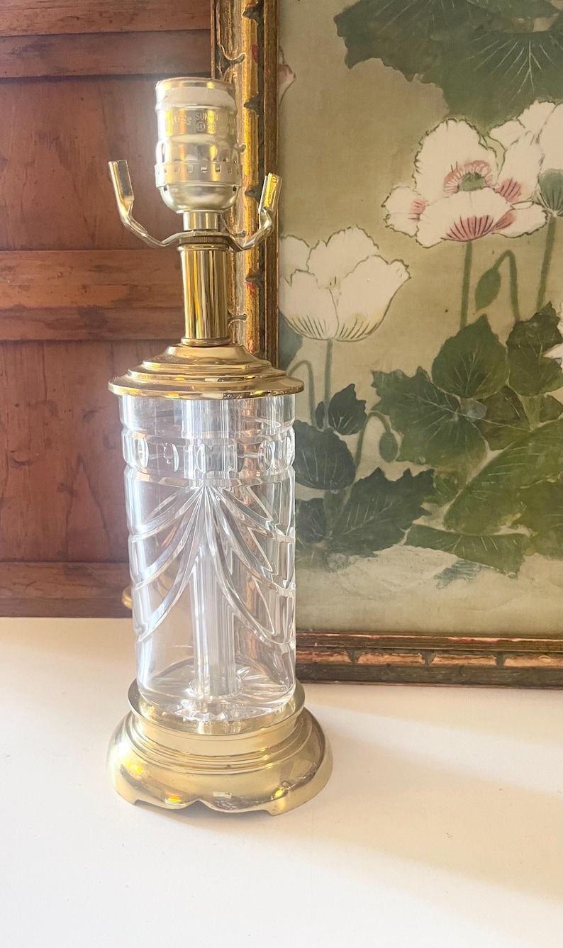 May include: A table lamp featuring a clear, cylindrical glass base with vertical etched detailing and a scalloped pattern. The lamp has a polished brass base, a matching brass neck, and a metal socket assembly at the top for a light bulb.