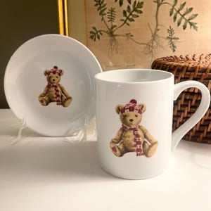 May include: A white ceramic mug and a matching white ceramic saucer, both featuring a centered illustration of a light brown teddy bear wearing a red and white plaid winter hat and scarf, with a small red heart detail on the bear's right foot.