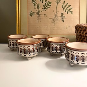 May include: A set of five small, round ceramic bowls, each featuring a smooth, light-colored interior and a brown rim. The exterior of each bowl is encased in a decorative, silver-toned metal sleeve with a repeating cutout pattern and three small feet.