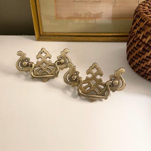 May include: Two brass drawer pulls featuring an ornate, openwork backplate with a symmetrical, lattice-like design and a curved, swinging bail handle. The metal has a polished, metallic gold finish with intricate scrollwork along the edges.