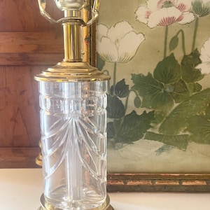 May include: A table lamp base featuring a clear, cylindrical glass body with vertical etched patterns, set upon a polished brass circular pedestal. The lamp includes a brass neck and a metal socket assembly at the top for a light bulb attachment.
