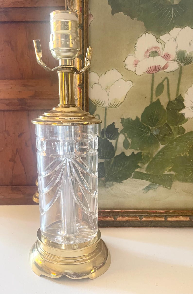 May include: A table lamp base featuring a clear, cylindrical glass body with vertical etched patterns, set upon a polished brass circular pedestal. The lamp includes a brass neck and a metal socket assembly at the top for a light bulb attachment.
