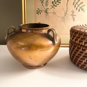May include: A vintage copper-toned metal vase with two curved handles. The vase has a rounded body and a dark interior. A woven basket is partially visible to the right, and a framed botanical print hangs in the background.