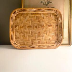 May include: A rectangular woven bamboo tray with rounded corners. The tray has a natural light brown color and a tightly woven pattern. The tray is sitting on a white surface, with a framed picture in the background.