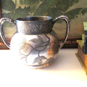 May include: A silver sugar bowl with an ornate floral design. The bowl has two handles and a round body. The design features leaves and flowers in shades of gold and silver.