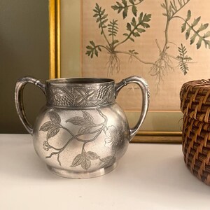 May include: A silver-toned metal sugar bowl featuring two curved handles and a rounded body. The exterior is decorated with embossed floral and vine motifs, including a band of grape leaves around the top rim and a central floral branch design.