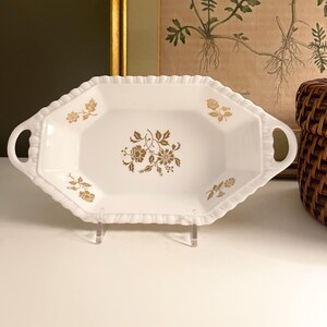May include: A white, octagonal serving tray with two integrated handles and a scalloped rim. The center and corners of the tray feature a gold-colored floral pattern. The tray is displayed on a clear stand, showcasing its symmetrical shape.