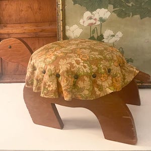 May include: A wooden turtle-shaped footstool with a floral patterned cushion. The cushion is a mix of orange, yellow, and green on a brown background. The wooden base is a medium brown color.