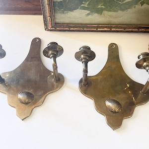 Puede incluir: Dos apliques de pared antiguos de latón. Cada aplique presenta una placa trasera decorativa con un diseño de concha y dos brazos curvos que sostienen copas para velas. Los apliques son de latón y tienen una pátina cálida y envejecida.