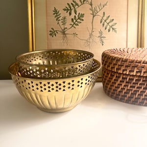 May include: Two gold-colored metal bowls, one nested inside the other, with decorative cut-out patterns. A woven basket with a lid sits to the right. A framed botanical print hangs in the background.
