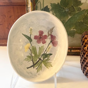May include: A small, off-white ceramic bowl with a floral design featuring red, yellow, and white flowers with green leaves. The bowl is displayed on a clear acrylic stand, showcasing its delicate details.