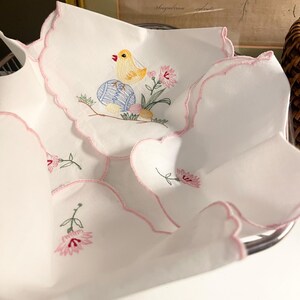 May include: White embroidered Easter napkins with scalloped edges. Each napkin features a yellow chick, a blue and white egg, and pink flowers. The napkins are neatly arranged in a metal basket, perfect for spring decor.