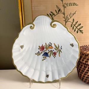 May include: A white ceramic dish shaped like a scallop shell with a scalloped edge and a green painted rim. The center features a hand-painted floral motif with red, blue, and yellow flowers and green leaves, with smaller floral accents on the sides.