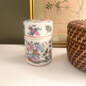 May include: A white ceramic jar with a lid, adorned with colorful floral and decorative patterns. The jar features a design of flowers in a vase and other Asian-inspired motifs. The jar is approximately 4 inches tall.