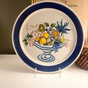 May include: A round ceramic plate with a blue rim and a central design of a fruit bowl. The bowl features a variety of fruits, including grapes, pears, peaches, and a pineapple. The plate has a crackle finish.