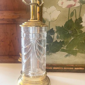May include: A table lamp base featuring a clear, cylindrical glass body with vertical cut-glass detailing and a polished brass metal base, neck, and socket assembly. The lamp has a standard light bulb socket at the top with two metal arms.