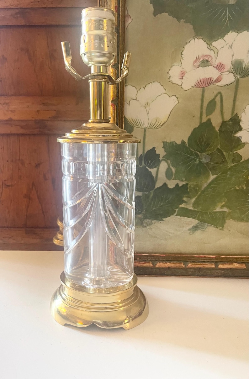 May include: A table lamp base featuring a clear, cylindrical glass body with vertical cut-glass detailing and a polished brass metal base, neck, and socket assembly. The lamp has a standard light bulb socket at the top with two metal arms.
