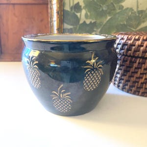 May include: A dark teal ceramic planter with gold trim and gold pineapple designs. The planter has a round shape and a wide opening. The background includes a wooden panel, a framed picture, and a wicker basket.