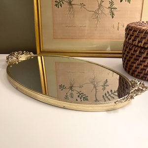 May include: An oval-shaped vanity tray with a mirrored base and a gold-toned metal frame. The tray features ornate, decorative metal handles on each end with intricate floral detailing and a textured, patterned edge along the perimeter.