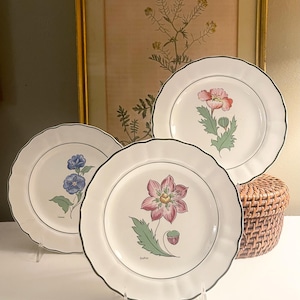 May include: Three white ceramic plates with scalloped edges and thin dark green rims. Each plate features a distinct botanical illustration of a flower with its name printed below in small black text, including a pink dahlia and blue flowers.