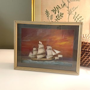 May include: Framed print of a tall ship sailing on the ocean under a sunset sky. The gold-colored frame surrounds the artwork, which features the text "H.M.S. Victory" at the bottom.