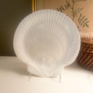 May include: A white, shell-shaped decorative dish. The dish has a ribbed texture and is displayed on a clear stand. The background includes a framed artwork and a woven basket, suggesting a coastal or nautical theme.