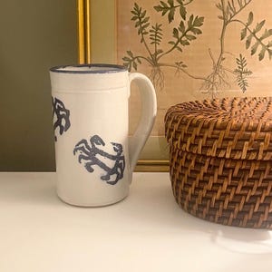 May include: A cylindrical ceramic mug with a white base and a blue rim. The exterior features a repeating pattern of dark blue crab illustrations. The mug has a single curved handle on the right side and a smooth, glossy finish throughout the surface.
