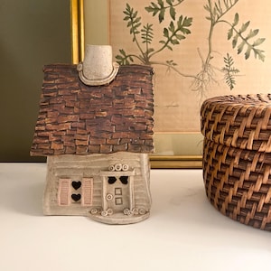 May include: A ceramic house-shaped candle holder with a textured brown shingled roof, a light beige facade, and a chimney. The front features a door with two small square windows and a window with pink shutters containing two heart-shaped cutouts.