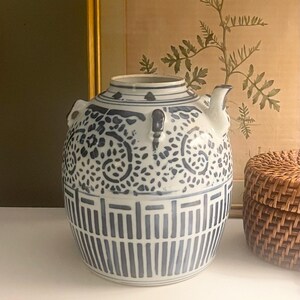 May include: A ceramic teapot featuring a white base with blue hand-painted patterns. The upper section displays intricate swirling floral motifs, while the lower portion is decorated with a series of vertical lines and rectangular geometric borders.