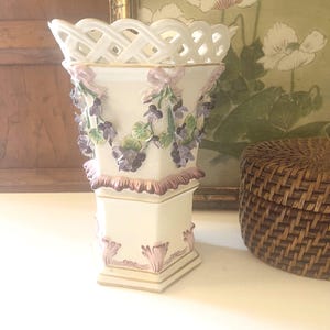May include: A white porcelain vase with a lattice top and floral embellishments. The vase features raised purple and green flowers and pink accents. The base has a square shape with gold trim, and the overall design is ornate and decorative.