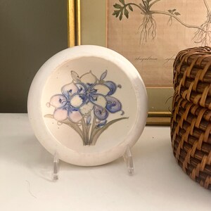 May include: A round, white ceramic dish featuring a hand-painted floral design in shades of blue, purple, and green. The dish is supported by a clear acrylic stand and displays a botanical illustration of a flower with multiple petals and stems.