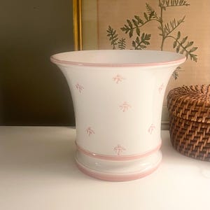 May include: A white ceramic vase with a flared rim and a rounded base, featuring a delicate pattern of small pink bows scattered across the exterior. The rim and the base are accented with a solid pink band, creating a clean and simple aesthetic.