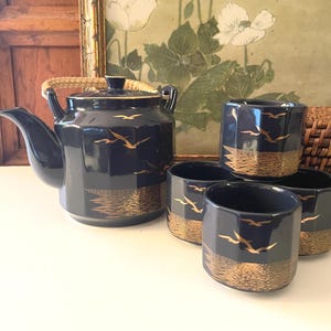 May include: A navy blue ceramic tea set with gold accents. The set includes a teapot and four cups. The cups and teapot feature a gold wave design and flying birds. The teapot has a gold rim and handle.