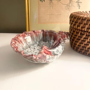 May include: A decorative ceramic bowl with a unique, organic shape. The bowl features a blend of red, white, and gray colors, with a textured interior. The rim has a wavy design. A woven basket is in the background.