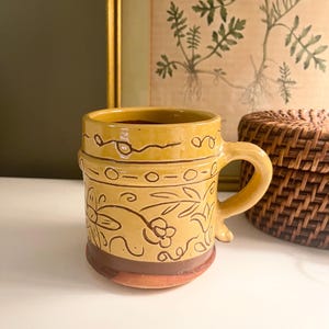 May include: A handmade ceramic mug with a yellow glaze and brown accents. The mug features a floral and leaf design, a handle, and a decorative rim. The mug is approximately 4 inches tall.