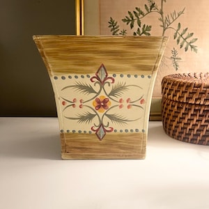 May include: A decorative wooden wastebasket with a light beige background and a floral design. The wastebasket has a rectangular shape with a flared top and is painted with a floral pattern in red, yellow, and gray.