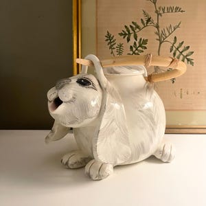May include: A white ceramic bunny teapot with gray brushstrokes. The teapot has a wooden handle and a lid. The bunny is in a sitting position with its mouth open. The teapot is on a white surface.