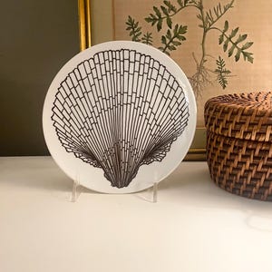 May include: A round white ceramic plate featuring a bold black line drawing of a large scallop shell centered on the surface. The plate is displayed upright on a clear plastic stand, showcasing the detailed radial ridges and fan-like shape.