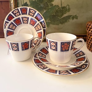 May include: A set of vintage teacups and saucers. The white porcelain cups and saucers feature a repeating pattern of blue squares, red flowers, and gold trim. The set includes two cups and saucers, and two additional saucers.