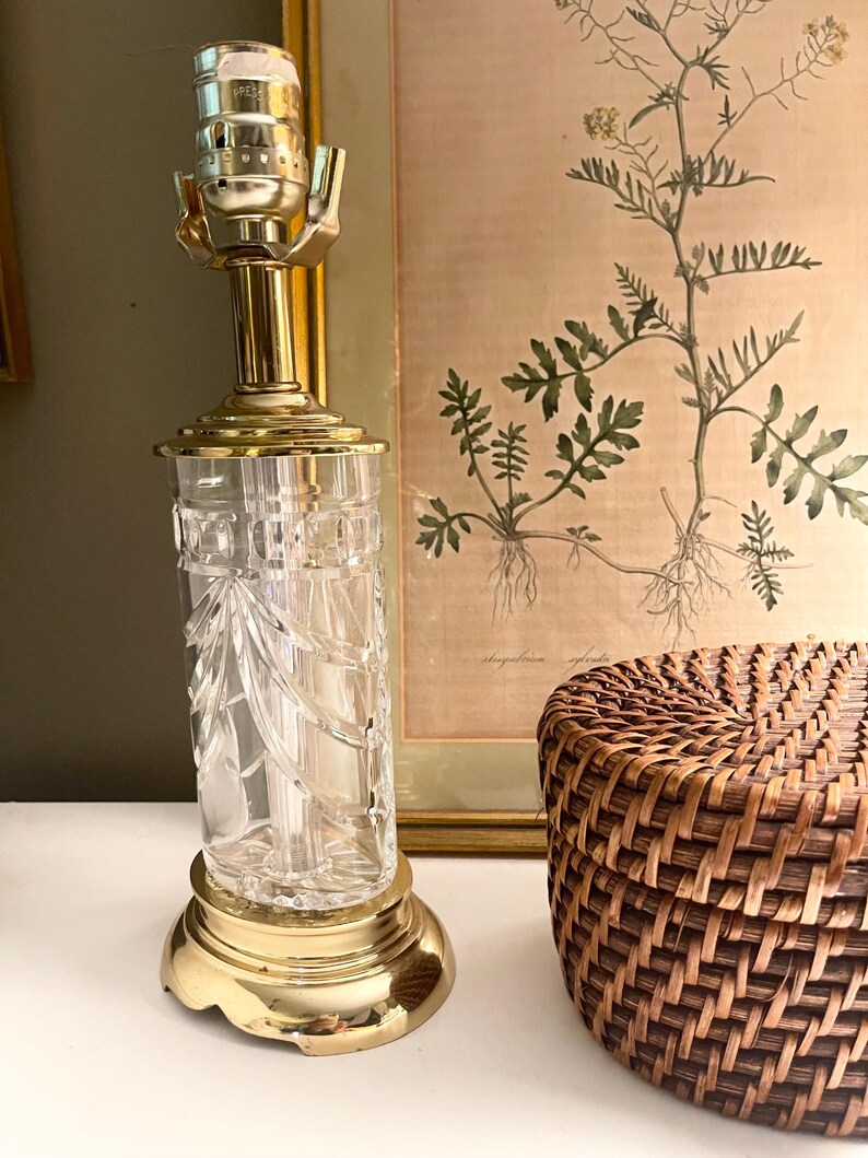 May include: A table lamp base featuring a clear, cylindrical glass body with vertical cut patterns. The lamp has a polished gold-tone metal base and a matching gold-tone metal socket assembly at the top, designed to hold a light bulb and shade.
