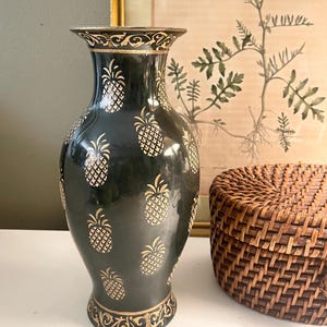 May include: A black vase with a glossy finish featuring a repeating gold pineapple pattern. The neck and base are decorated with intricate gold scrollwork. The vase has a flared rim and a tapered body, standing upright with a smooth, reflective surface.