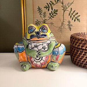 May include: A ceramic frog figurine featuring a vibrant hand-painted design with green, orange, blue, and black patterns. The frog has large eyes, a wide mouth, and sits in a seated position with its front limbs resting on its belly.