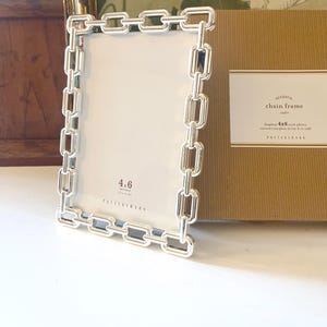 May include: A silver chain frame for a 4x6 inch photo. The frame is rectangular with a chain link design around the edges. The frame is next to a brown box with the text "chain frame".