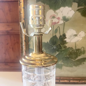 May include: A clear glass lamp base featuring a faceted geometric pattern and a polished brass neck. The top includes a silver-toned metal light socket with two extending arms for shade attachment, showcasing a reflective and transparent design.